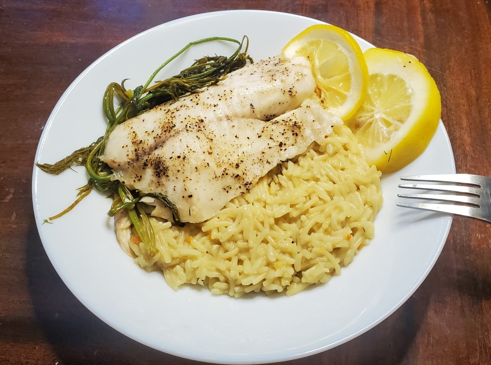 Plate with a lightly seasoned tilapia fillet on yellow rice with baked dill & lemon on the sides as garnish.